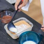 Photographie d'une louche noire tupperware en plastique utilisée par un chef pour verser un liquide blanc dans un récipient transparent contenant des ingrédients de dessert. Pichet bleu et bol de chocolat fondu présents."